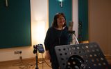 A woman stands in front of a microphone in a recording booth. She is smiling.