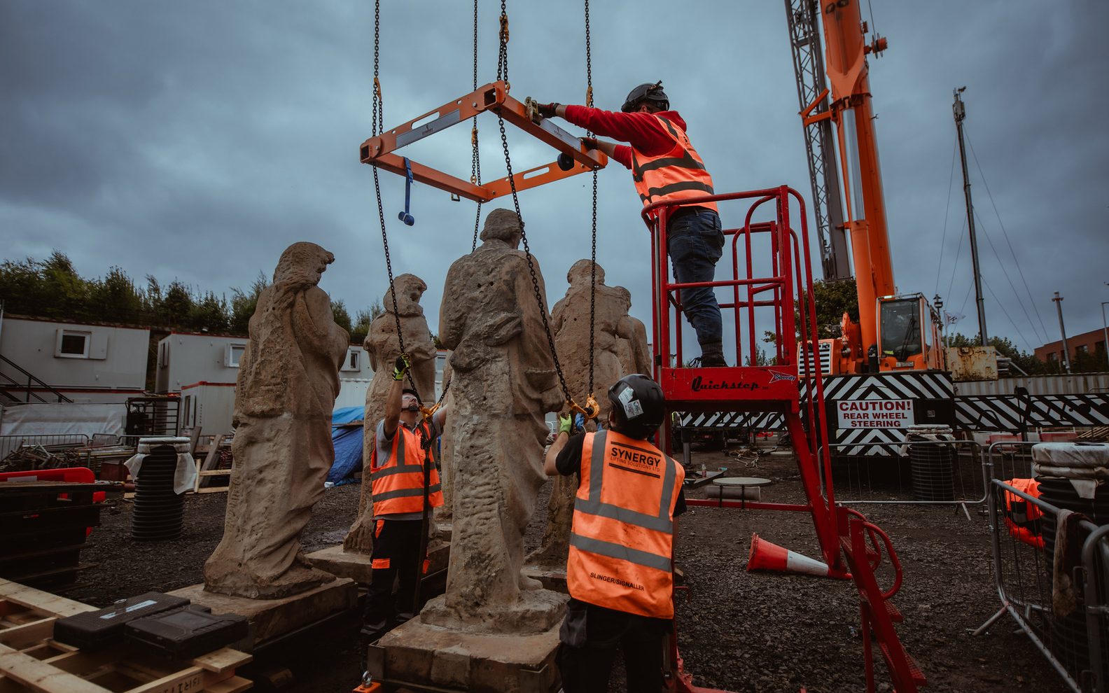 The statues preparing to be lifted onto the roof. People in hi-viz vests are securing the crane onto one of the statues.