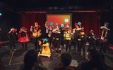 A large group of people standing on a stage. They are all wearing large hats, red clown noses and holding colourful props. In the foreground, you can see the backs of an audience .