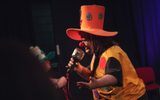 A man dressed as a clown. He is speaking into a large prop microphone and looks like he is telling the audience a joke.