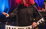 A woman standing on stage. She has a large pink hat and is playing an oversized prop keyboard.