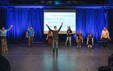 A group of young people performing on stage. One man is standing in the centre with his arms raised in the air. A sign behind him says ' I like this song because it's Scottish and I'm Scottish. Being Scottish is important to me.'