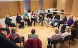 A play reading session. A group of older adults sit in a circle, holding books.