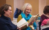 A play reading session. A woman with glasses and a bright blue scarf smiles while reading from a book.