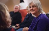 A woman in glasses smiles and listens to someone off-camera.