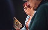 A play reading session. A woman with glasses and short red hair reads from a copy of the play 