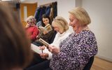 A play reading session. Four women sit in a row and read from copies of the same play. They are laughing.