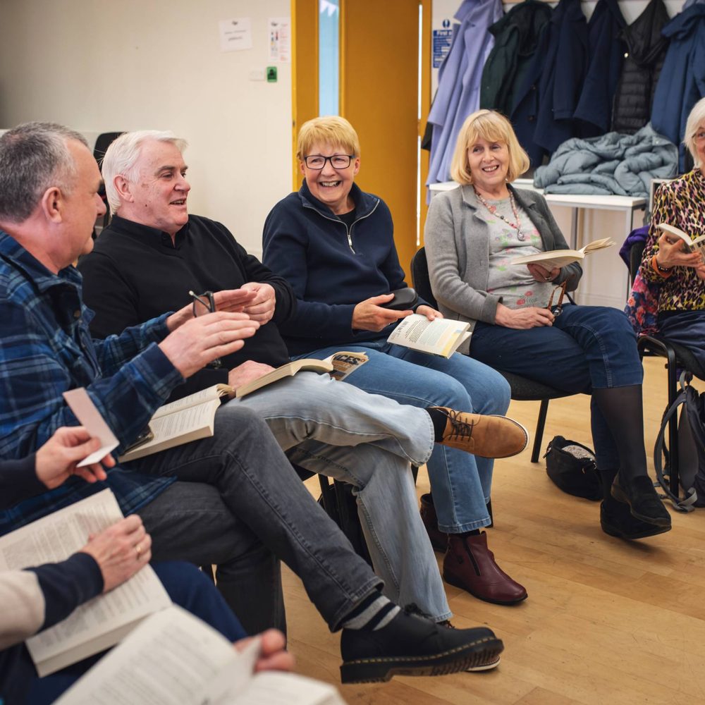A play reading session. A group of older adults sitting in a circle, holding copies of the same book and laughing.