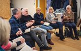 A play reading session. A group of older adults sitting in a circle, holding copies of the same book and laughing.
