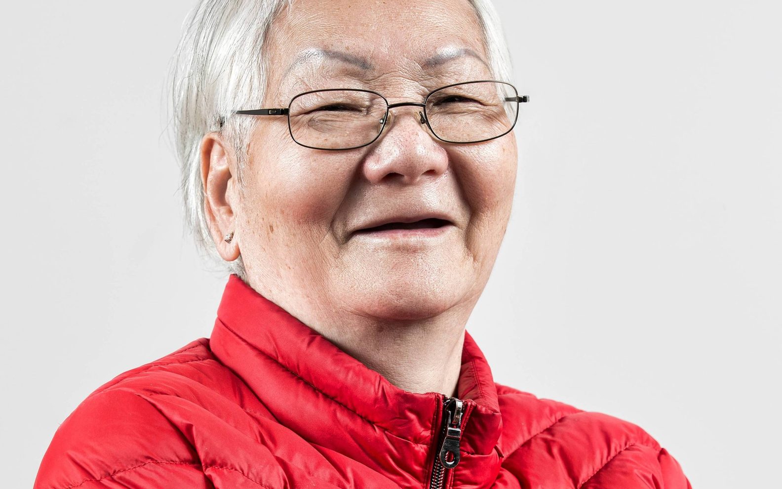 An older Asian woman with white hair smiles at the camera. She is wearing a bright red puffer jacket.