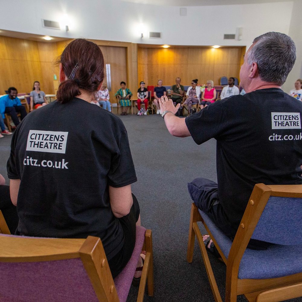 A large group of people sitting in a circle. The two people in the centre of the image are wearing Citizens Theatre t-shirts.