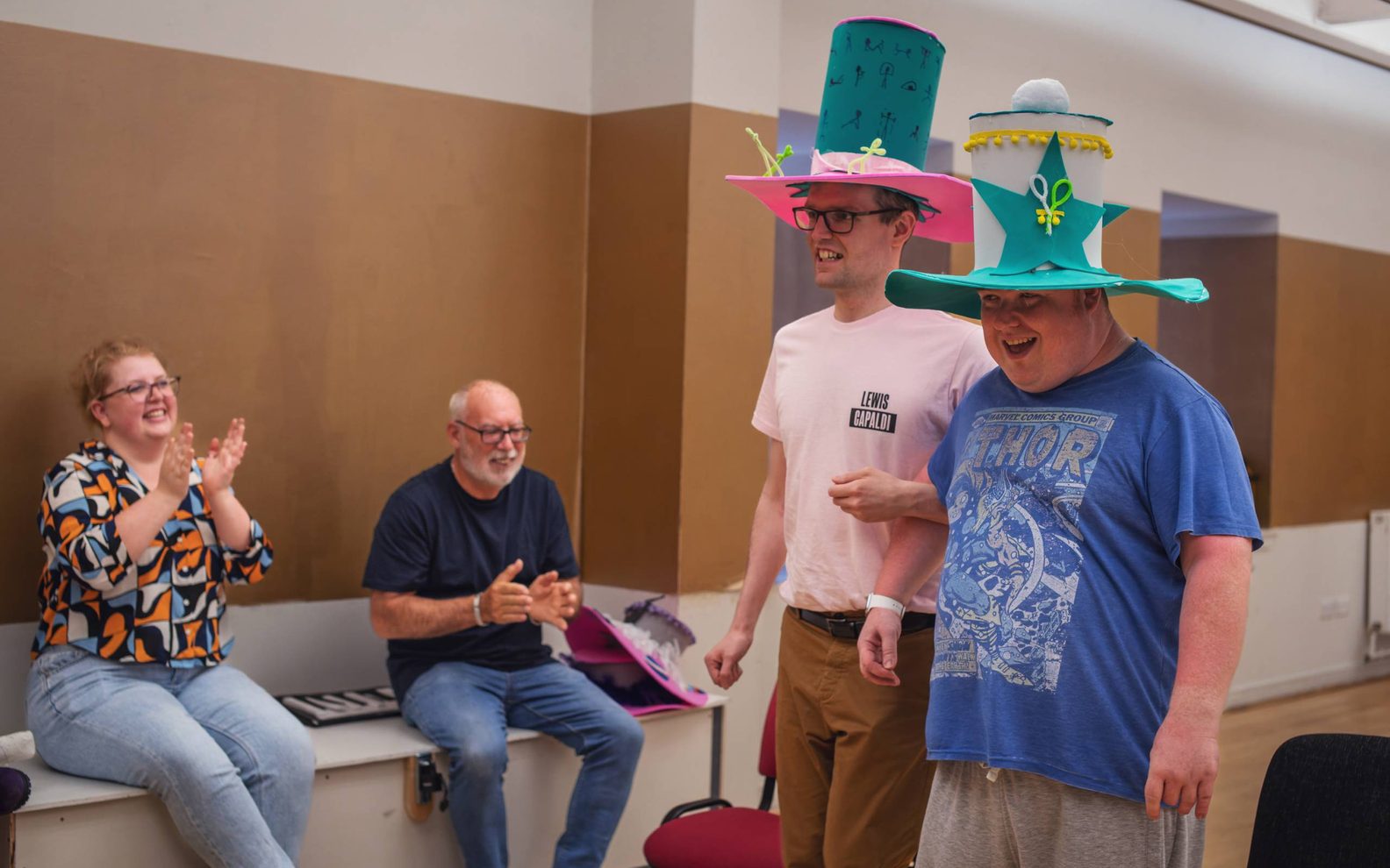A drama workshop for learning disabled adults. Two young men wearing large brightly coloured hats are walking arm in arm.A man and a woman are sitting on a bench nearby clapping.