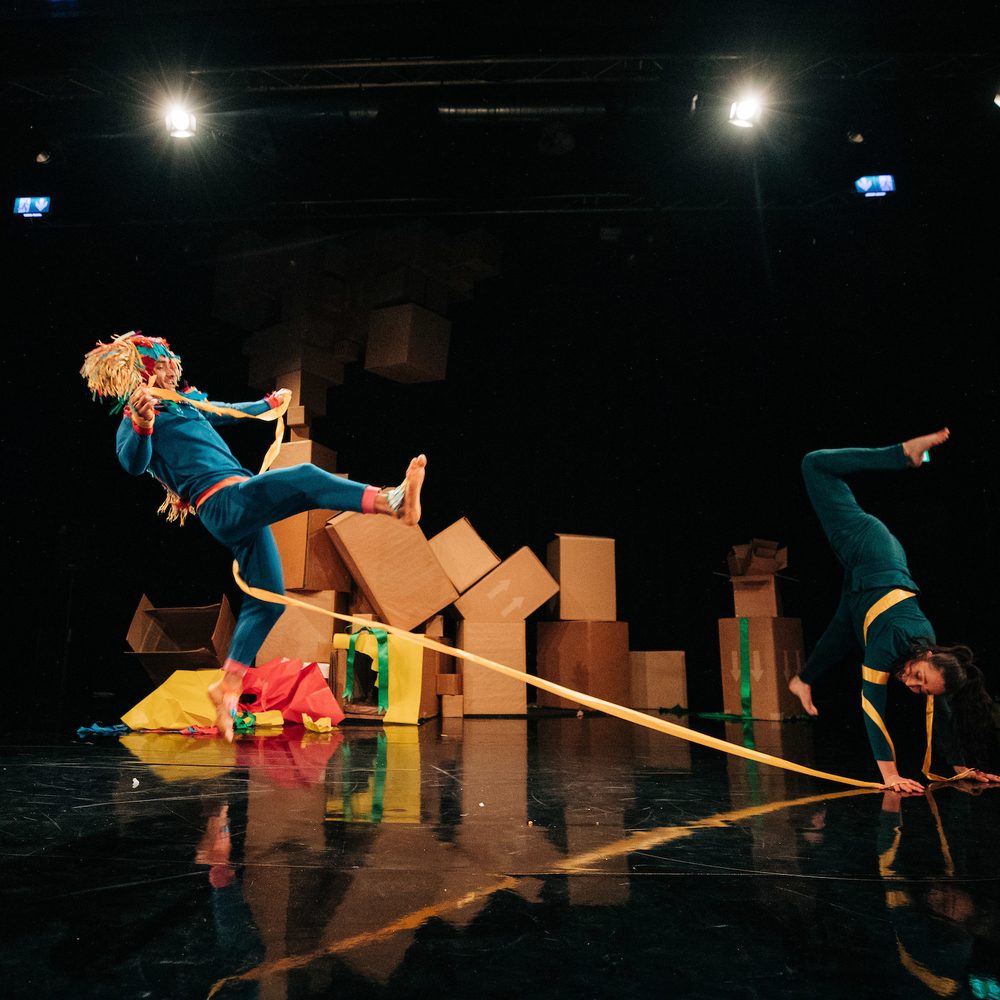 Two performers on stage. One is leaping and kicking their legs in the air. The other is doing a handstand. They both have a long piece of ribbon wrapped around them, and there are piles of cardboard boxes on the stage behind them.