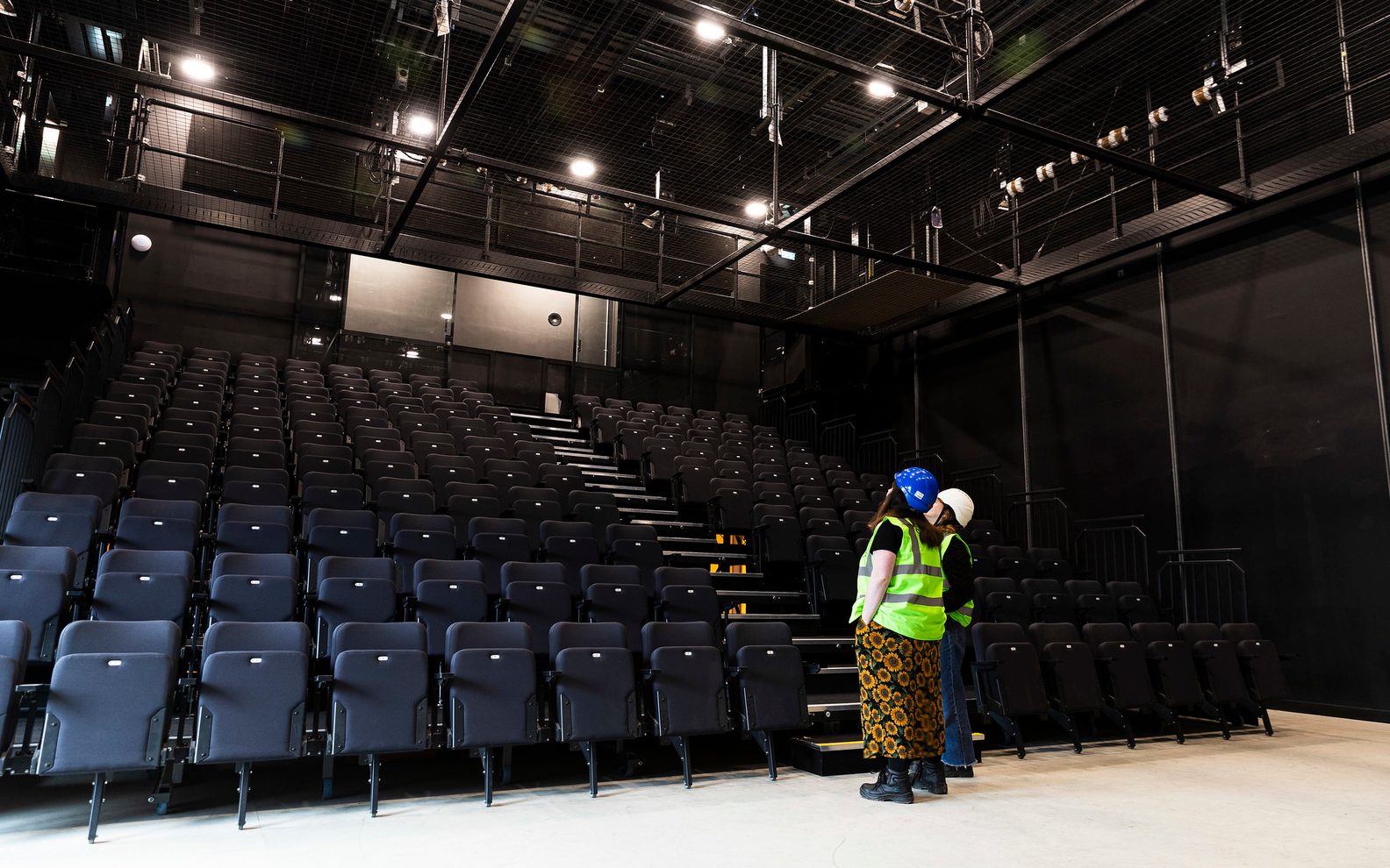 Two people standing in a a studio theatre space looking up towards the top of the seating bank. They are wearing high vis vests and hard hats