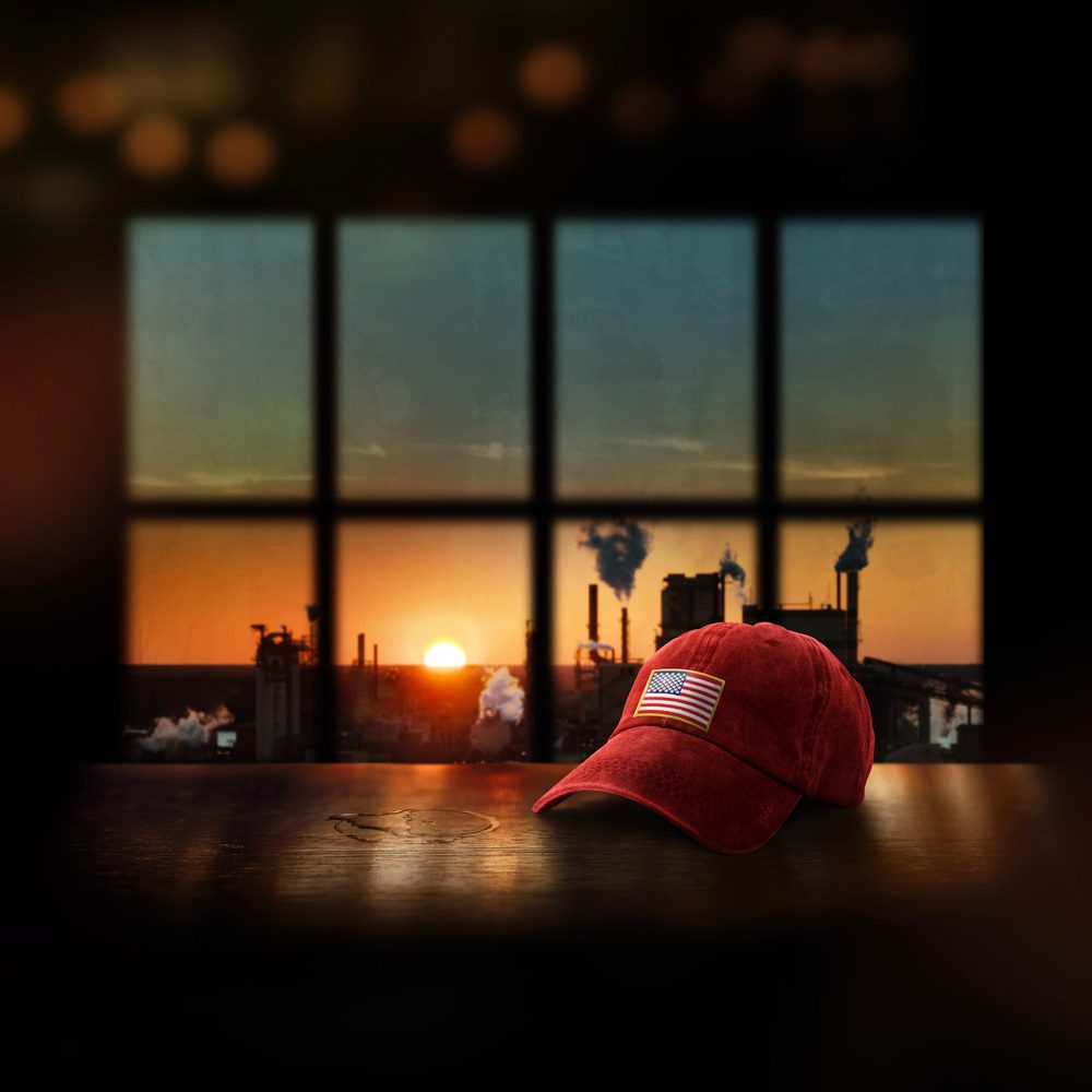 An American cap sitting on a table. Behind is a window where you can see a sunset and indistrial chimney.