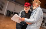 Two men are standing close each other. They are holding scripts and laughing.