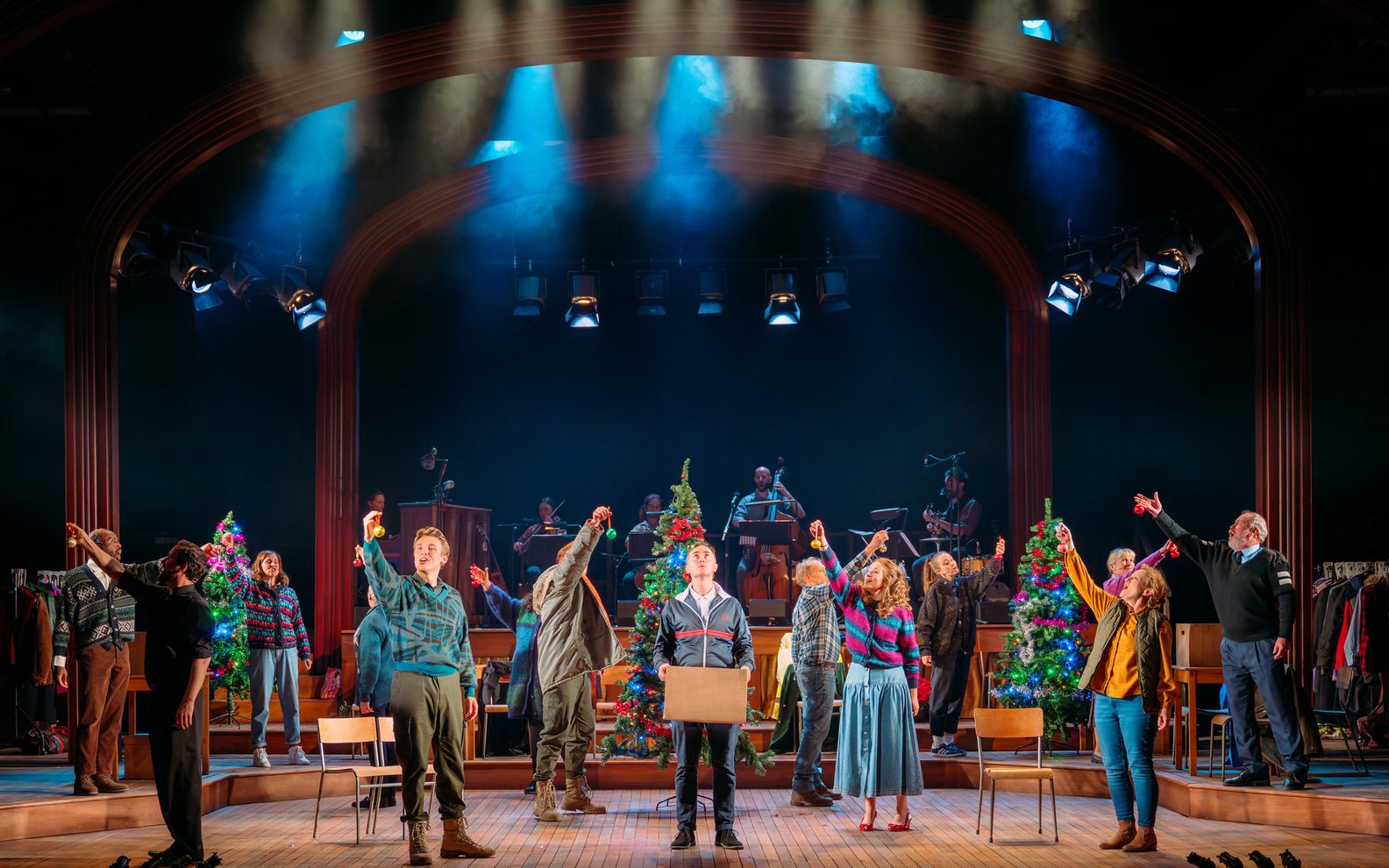 14 people on a stage stand holding Christmas ornaments in front of them, facing in different directions, and one man centre stage holds a cardboard box. A Christmas tree is lit in the middle of the stage.