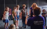 A group of children watching an adult who is facing away from the camera. The adult is sat down and has Citizens Theatre on the back of their purple t-shirt.