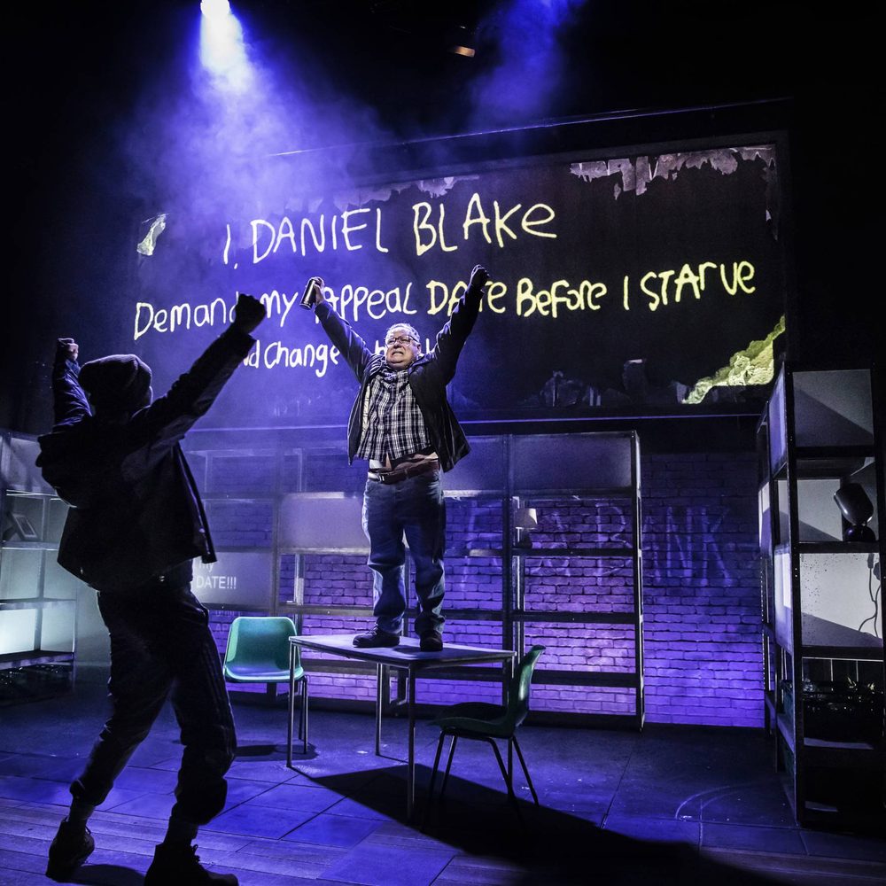 A man stand on a chair with another man cheering in front of him in shadow, with white spray paint in the background reading 'I, Daniel Blake'.