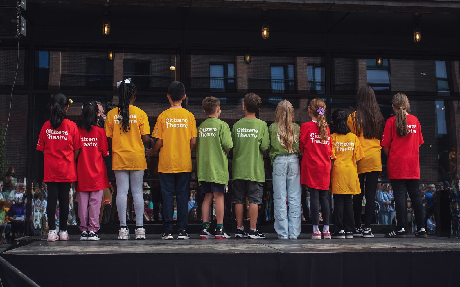11 children wearing red, yellow and green t-shirts facing away from the camera. Each has 'Citizens Theatre' on the back of their t-shirts.