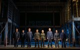 Eight actors all dressed in dark boiler suits are standing in a row.