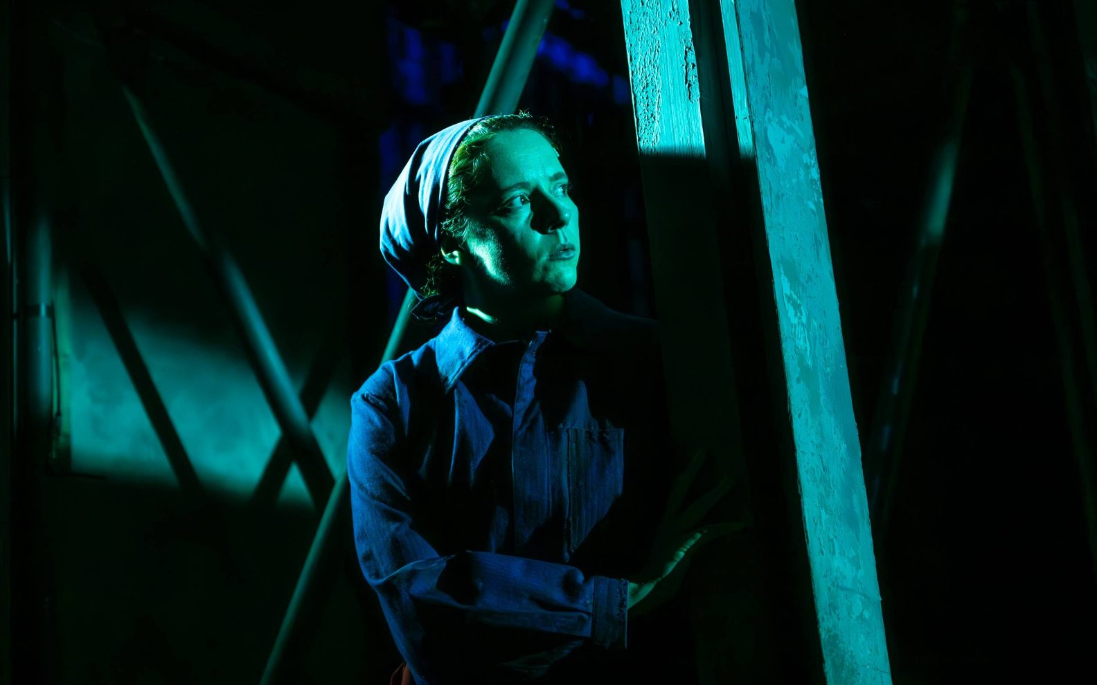 A female actor wearing a blue boiler suit is lurking in the shadows.