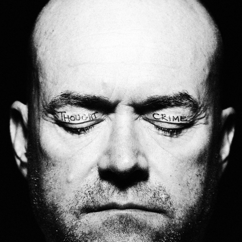 A close up photo of a man's face. He has his eyes closed and the words 'thought' and 'crime' are tattooed on his eyelids.