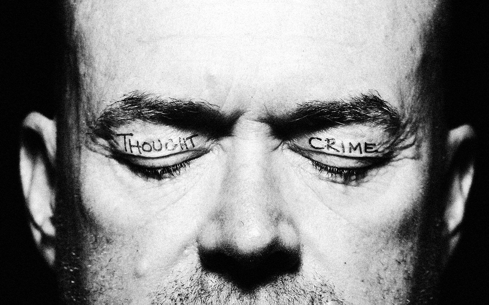 A close up photo of a man's face. He has his eyes closed and the words 'thought' and 'crime' are tattooed on his eyelids.