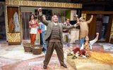 East is East cast members cheering with their arms raised. The set looks like a 1970s style kitchen and there are all wearing 1970s style clothing.