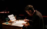 A performer on stage. The man is sitting at a desk writing. On the desk is an egg in a egg cup, an hourglass timer and a mobile phone.