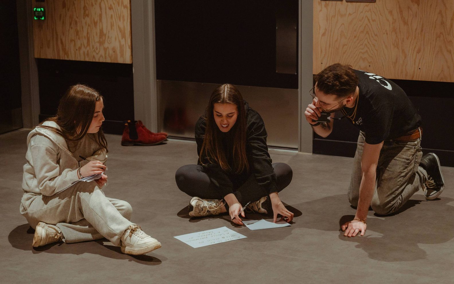Three teenagers are sitting on the floor. They are all looking at two sheets of paper.