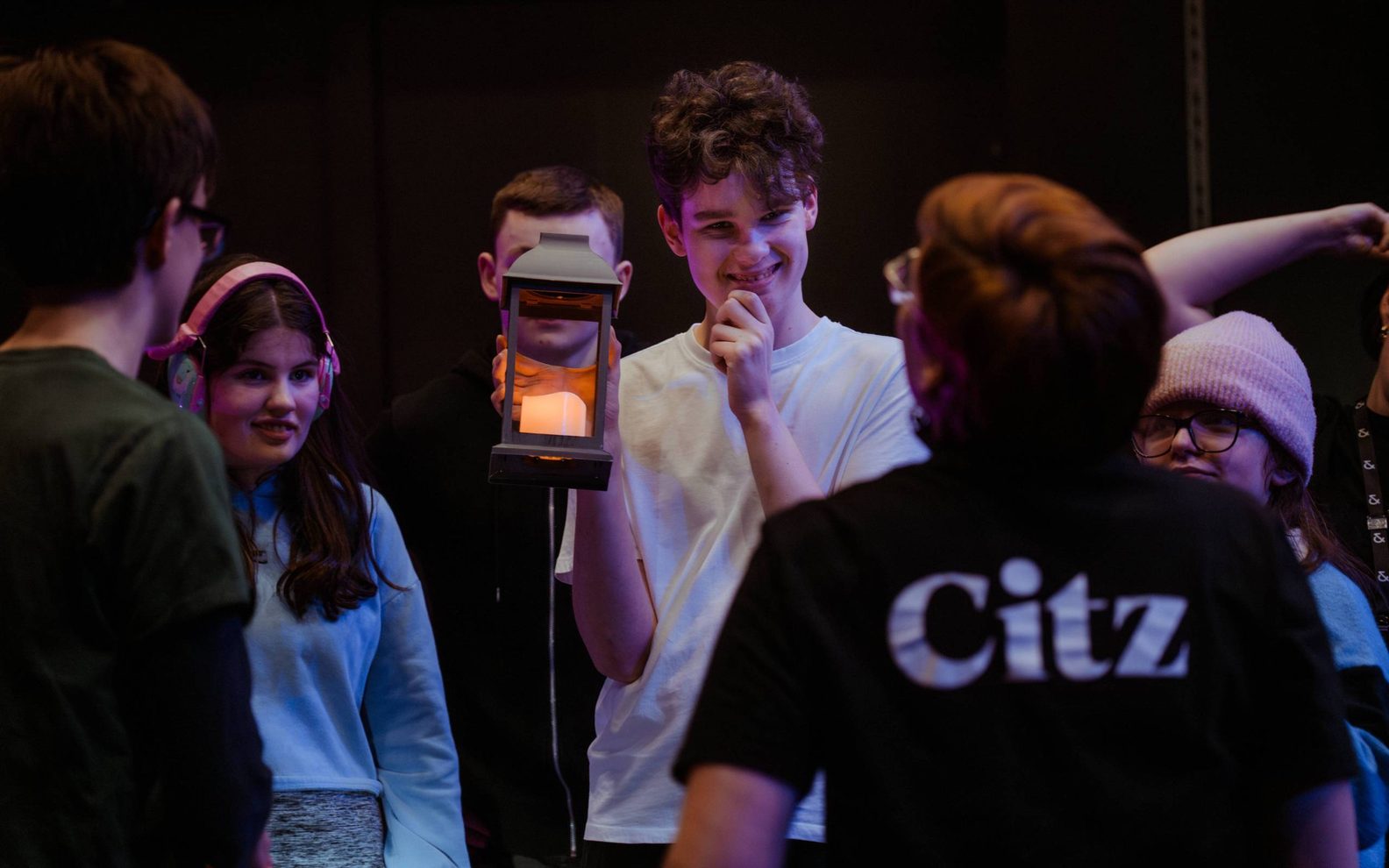 A group of young people are standing looking at an adult wearing a Citizens Theatre t-shirt. One young person is holding up a lamp.