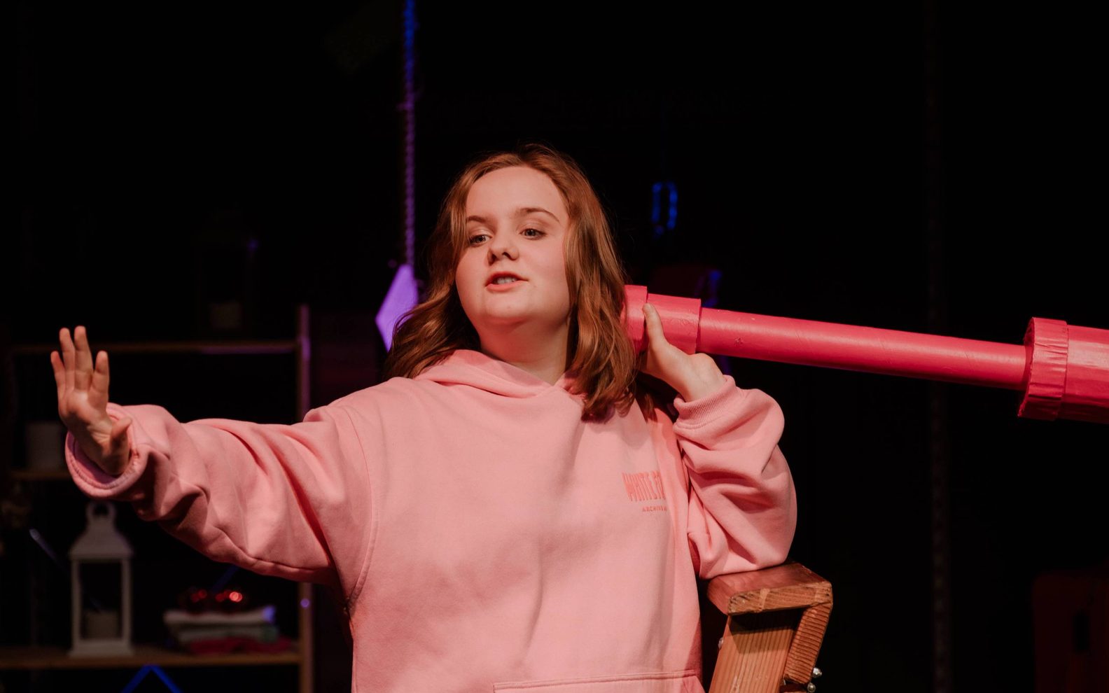 A girl in a pink hooded top is standing. She has a long pink tube held up to her ear and her arm is stretched out in front of her.