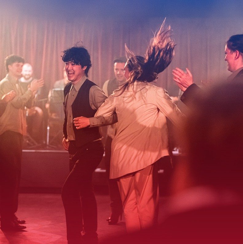 A group of people are dancing energetically. Two people in the middle are holding hands. There is a red lisght across the room.
