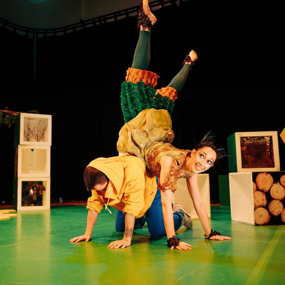 Two performers are dressed in brightly coloured clothes. One is kneeling on all fours on the group. The other is doing a handstand against them.