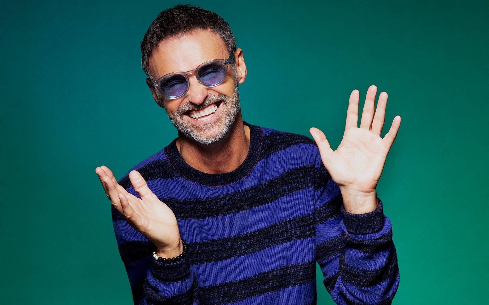 A man, Marti Pellow, is sitting in front of a green background. He is wearing a blue and black striped jumper, blue tinted glasses and is smiling.